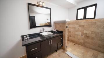 Un baño con encimera negra, lavamanos blanco, mueble color café oscuro, espejo y zona de ducha con baldosas beige.