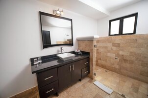 Un baño con encimera negra, lavamanos blanco, mueble color café oscuro, espejo y zona de ducha con baldosas beige.