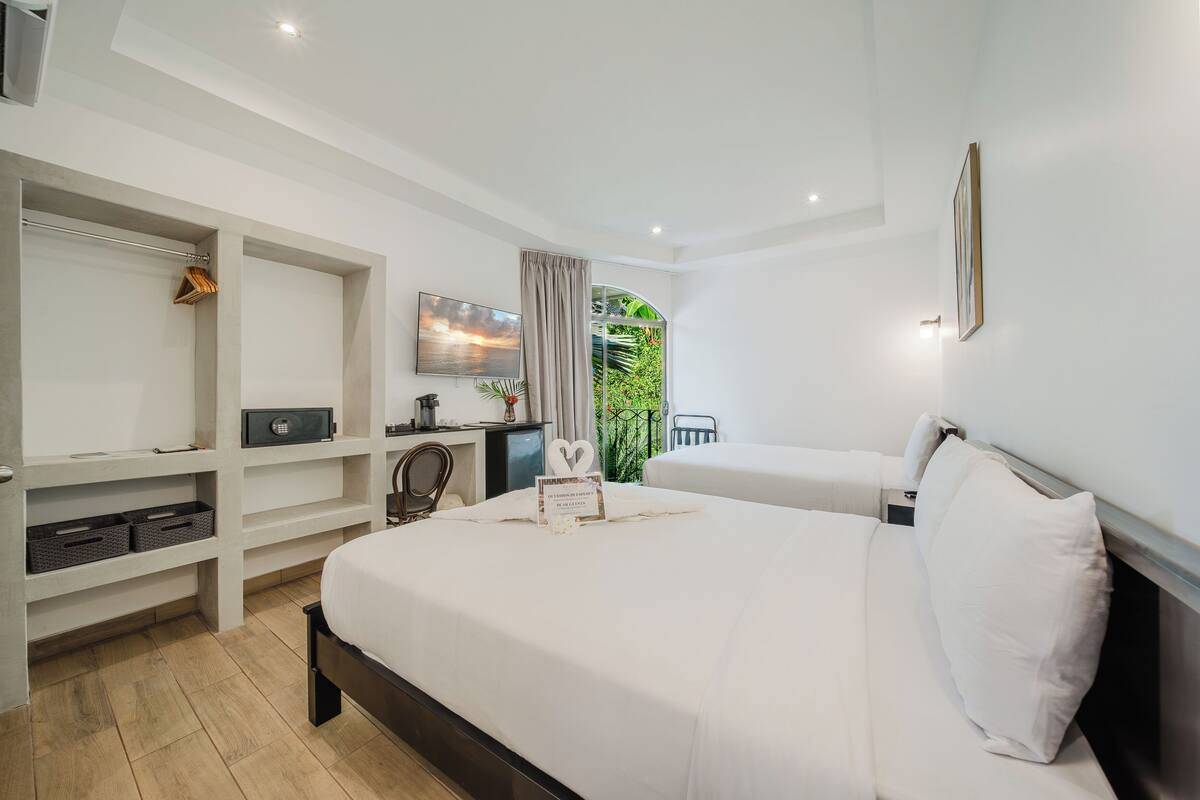 A hotel room with a bed, a desk, a chair, a TV, and a balcony with a view of greenery.
