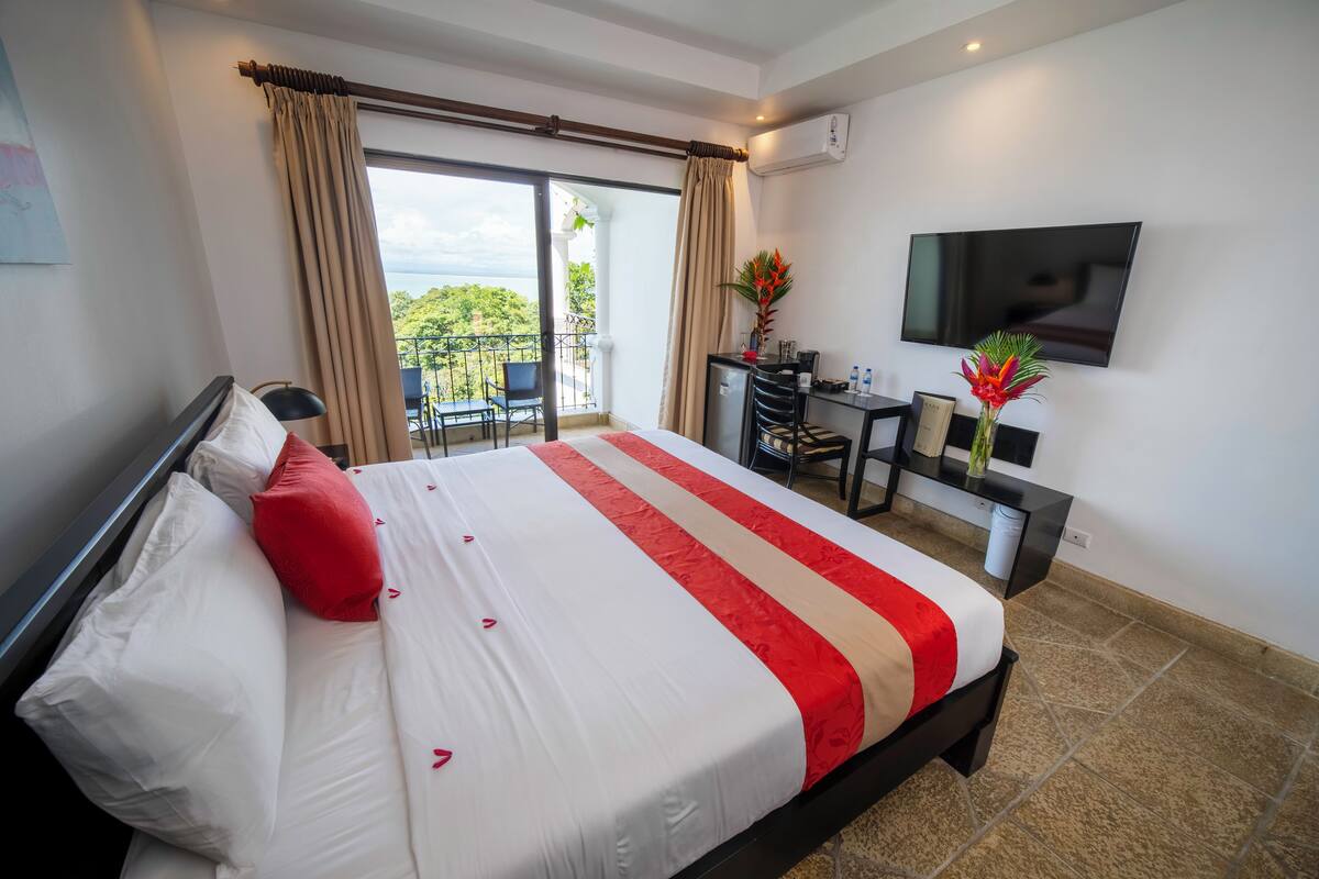 A hotel room with a bed, a desk, a TV, and a balcony with a view.