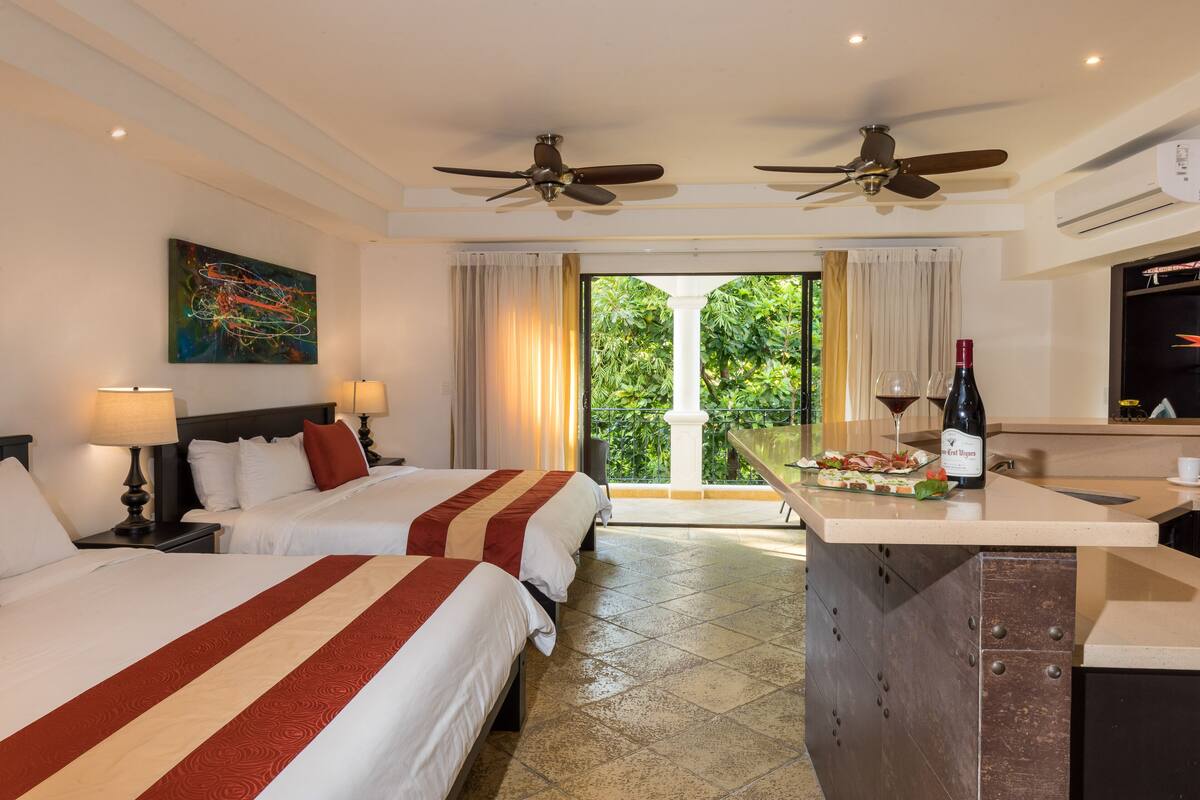A hotel room with two beds, a dining area with a bottle of wine, and a balcony with a view of greenery.