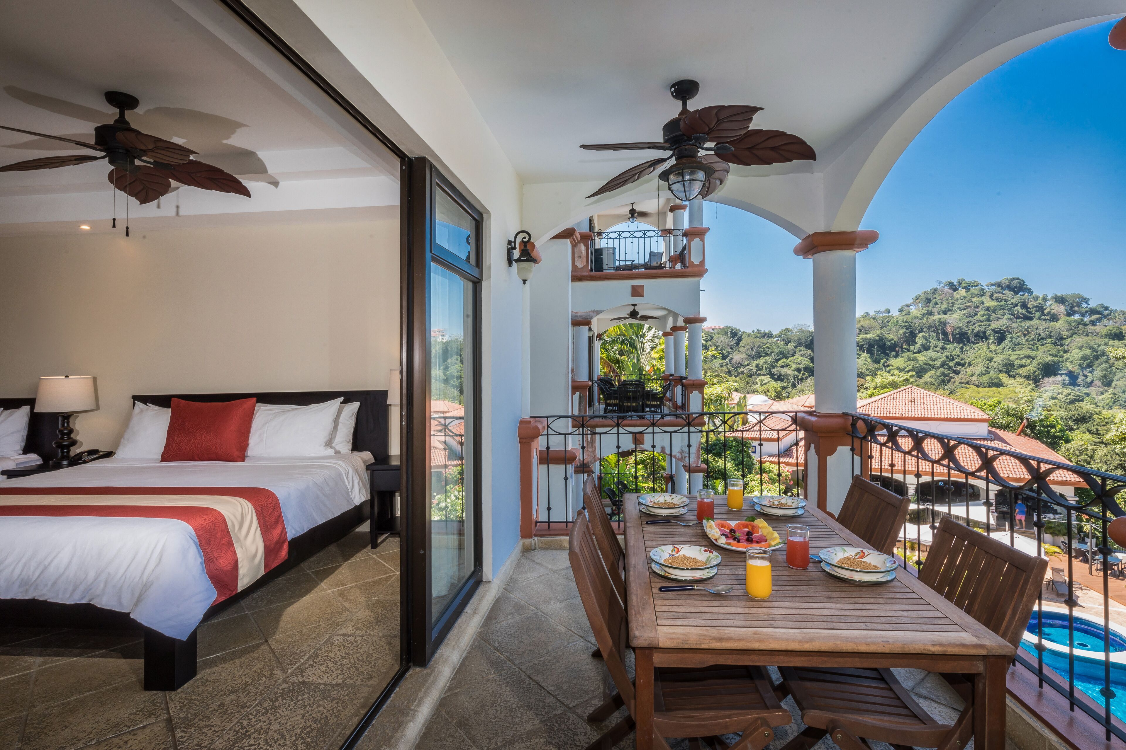 A hotel room with a balcony, a bed, a dining table with breakfast, and a pool view.