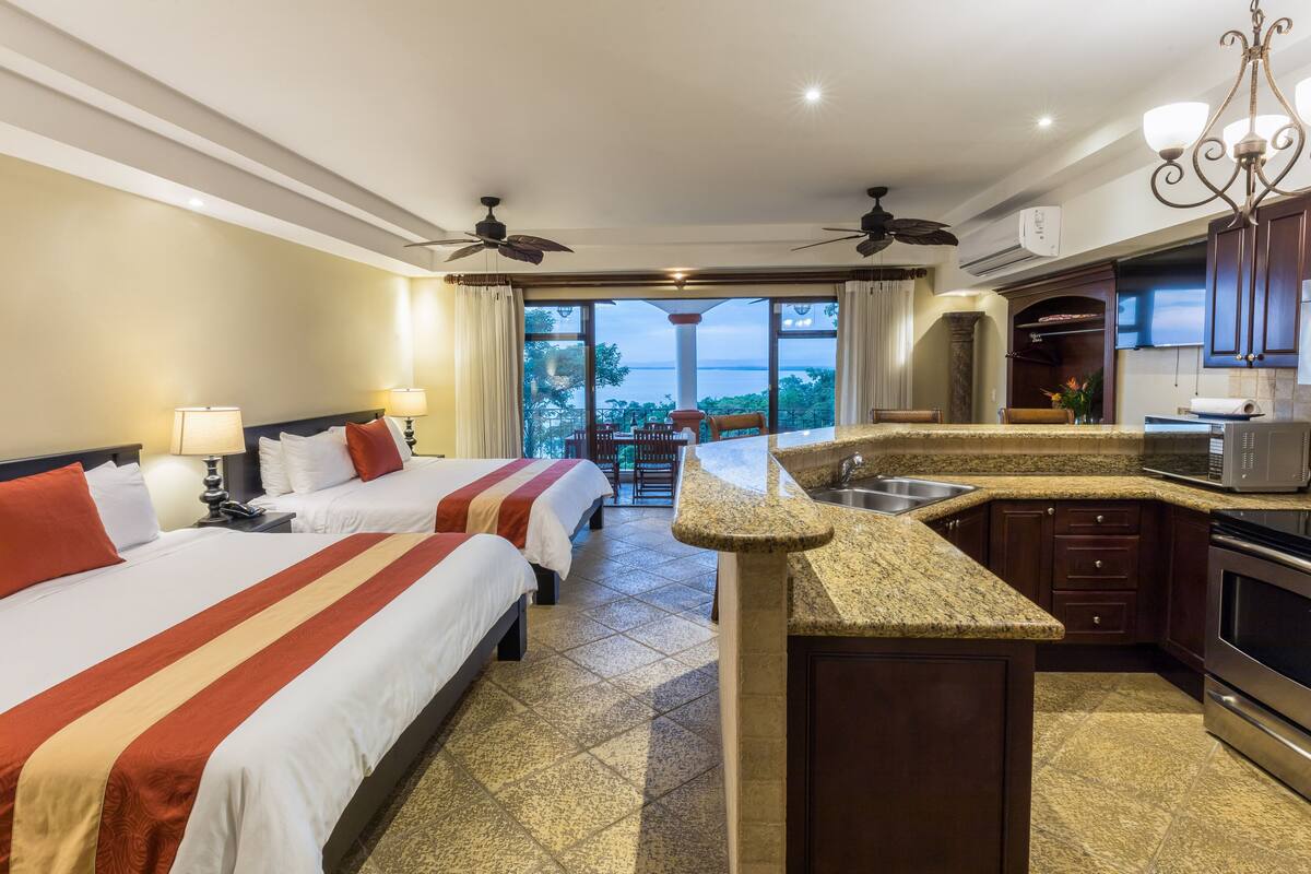 A hotel room with two beds, a kitchenette, and a balcony with a view of the sea.