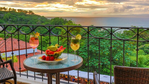 A balcony with a table set for two, featuring a fruit bowl and two drinks, overlooking a lush landscape and the sea. - Shana by the Beach Manuel Antonio (Quepos)