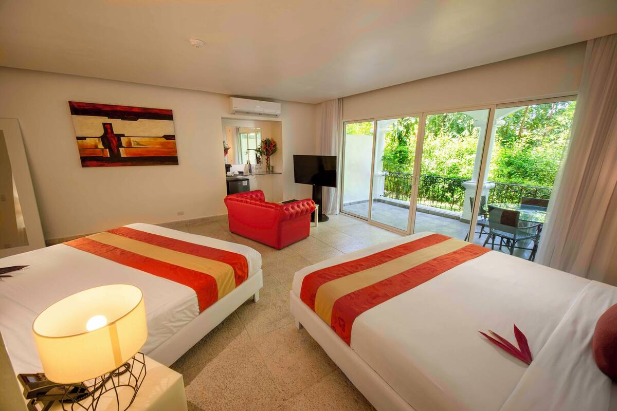 A hotel room with two beds, a red sofa, a TV, and a balcony with a view of greenery.