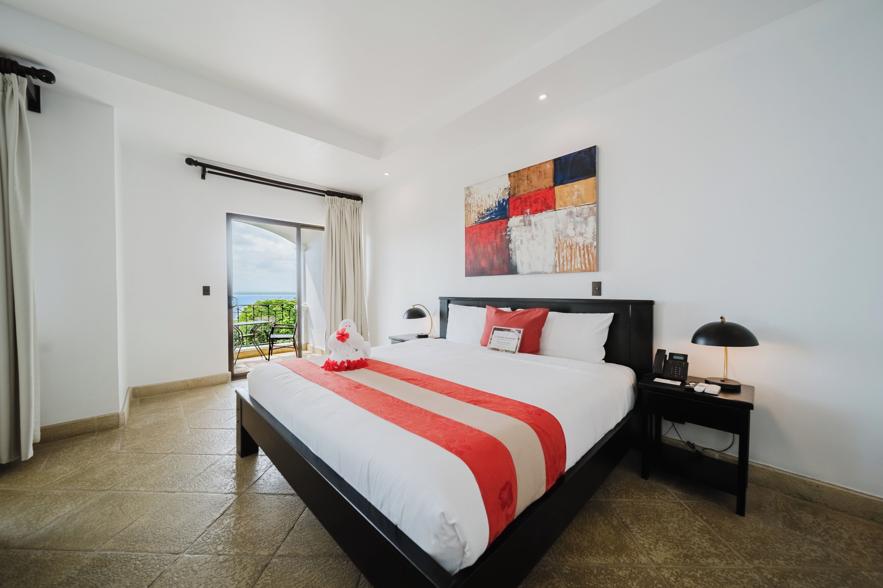 A hotel room with a large bed, a nightstand with a lamp, a painting on the wall, and a view of the outdoors.