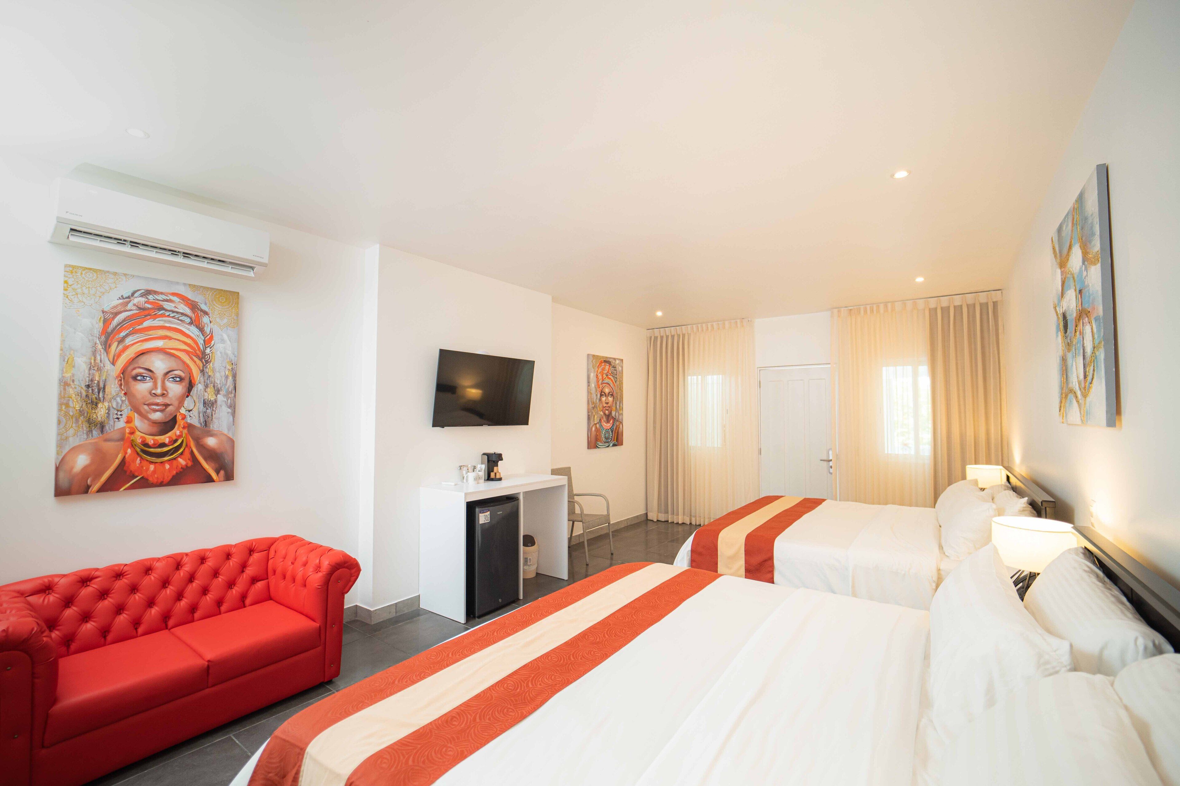 Une chambre d’hôtel avec deux lits, un canapé rouge, un bureau avec une télévision et des œuvres d’art accrochées aux murs.