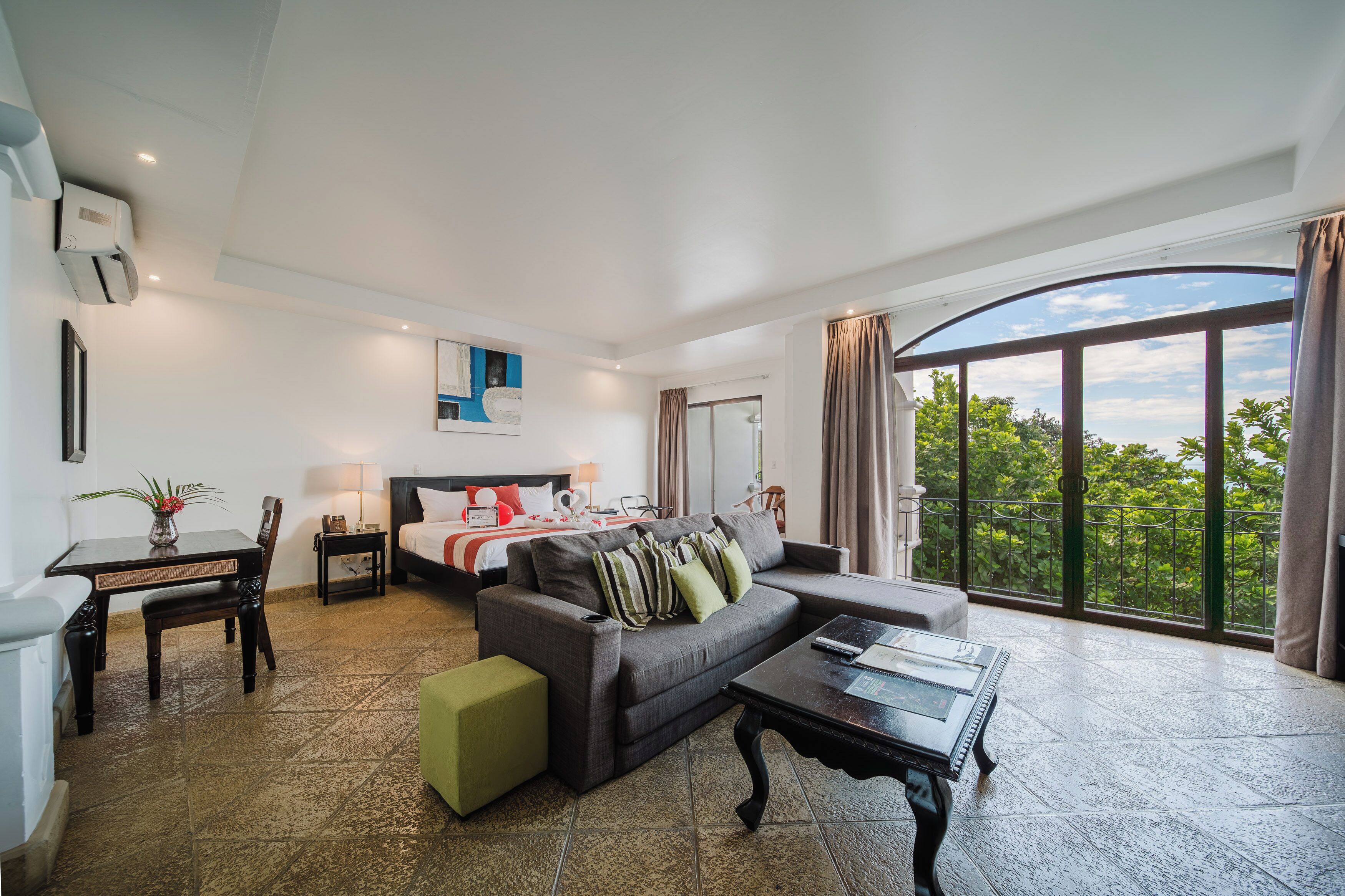 A hotel room with a bed, sofa, dining table, and a large window with a view.