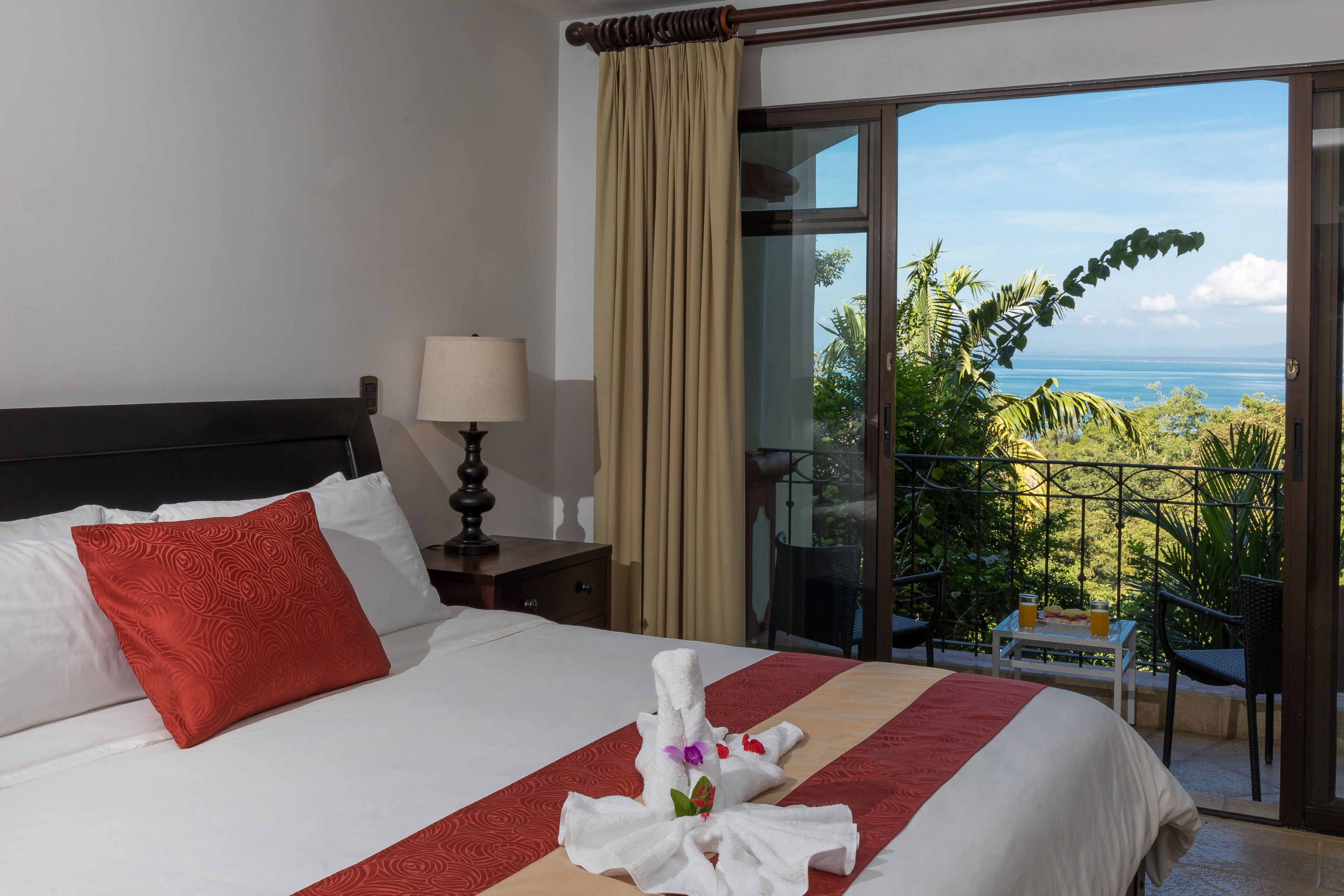 A hotel room with a bed, a nightstand, a lamp, a balcony with a view of the ocean, and a table with a tray of breakfast items.