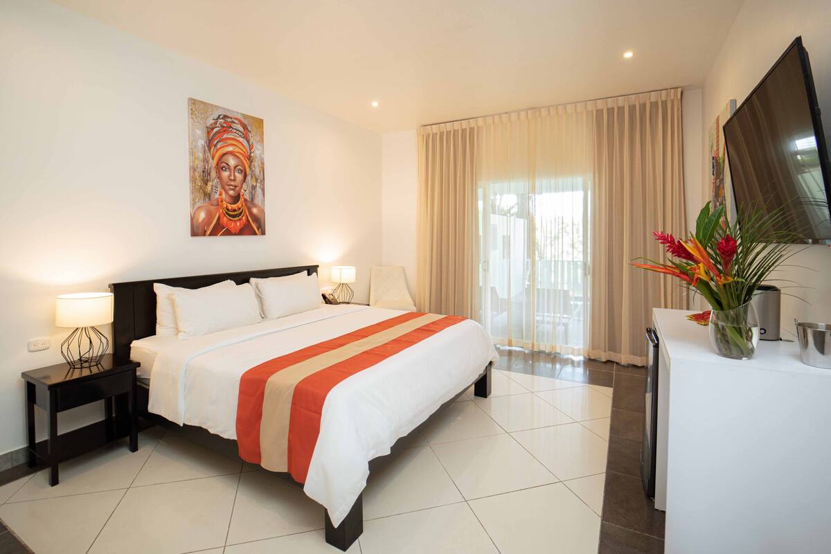 A hotel room with a large bed, bedside tables with lamps, a flat-screen TV, a desk with a vase of flowers, and a large window with curtains.