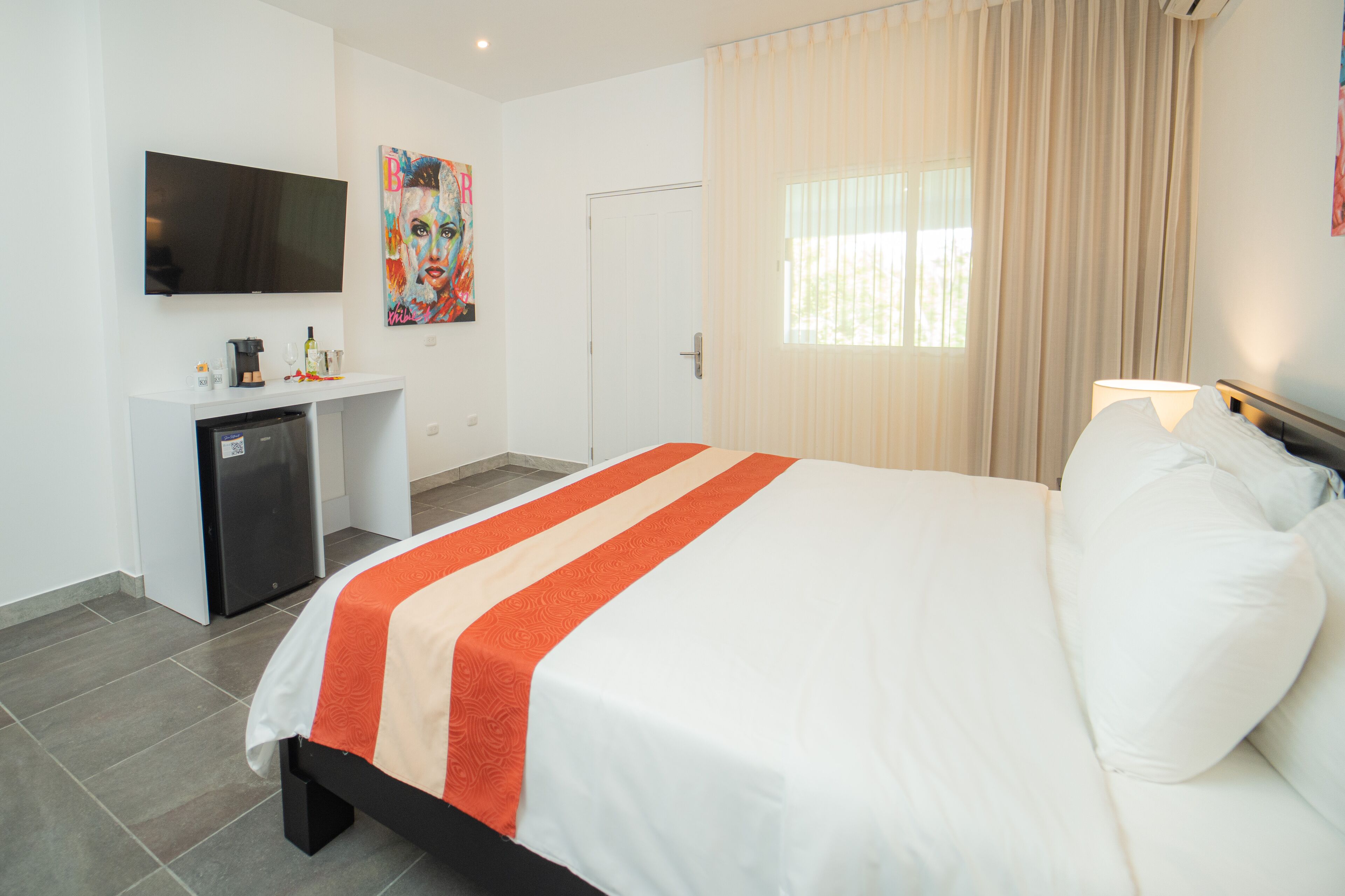 A hotel room with a bed, a TV mounted on the wall, a desk with a refrigerator, and a window with curtains.