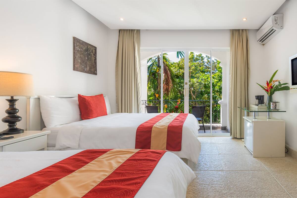 A hotel room with two beds, a desk, a TV, and a balcony with a view of greenery.
