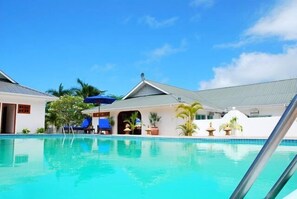 A swimming pool with lounge chairs and umbrellas, surrounded by buildings with a tropical design. - The Britannia Hotel (Praslin Island)