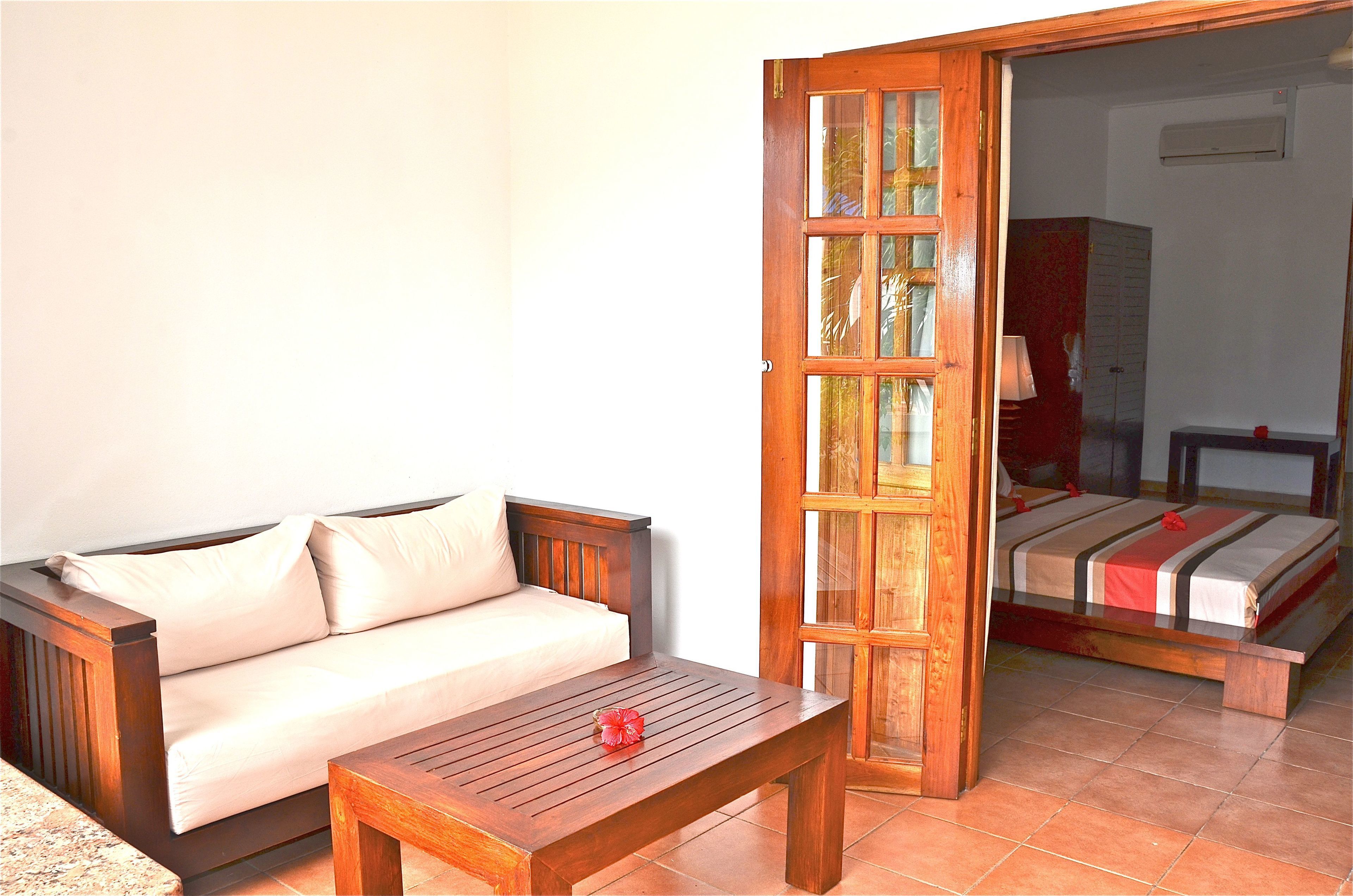 Quarto com sofá de madeira, uma mesa de madeira com uma flor vermelha e uma porta aberta para uma cama com colcha listrada.