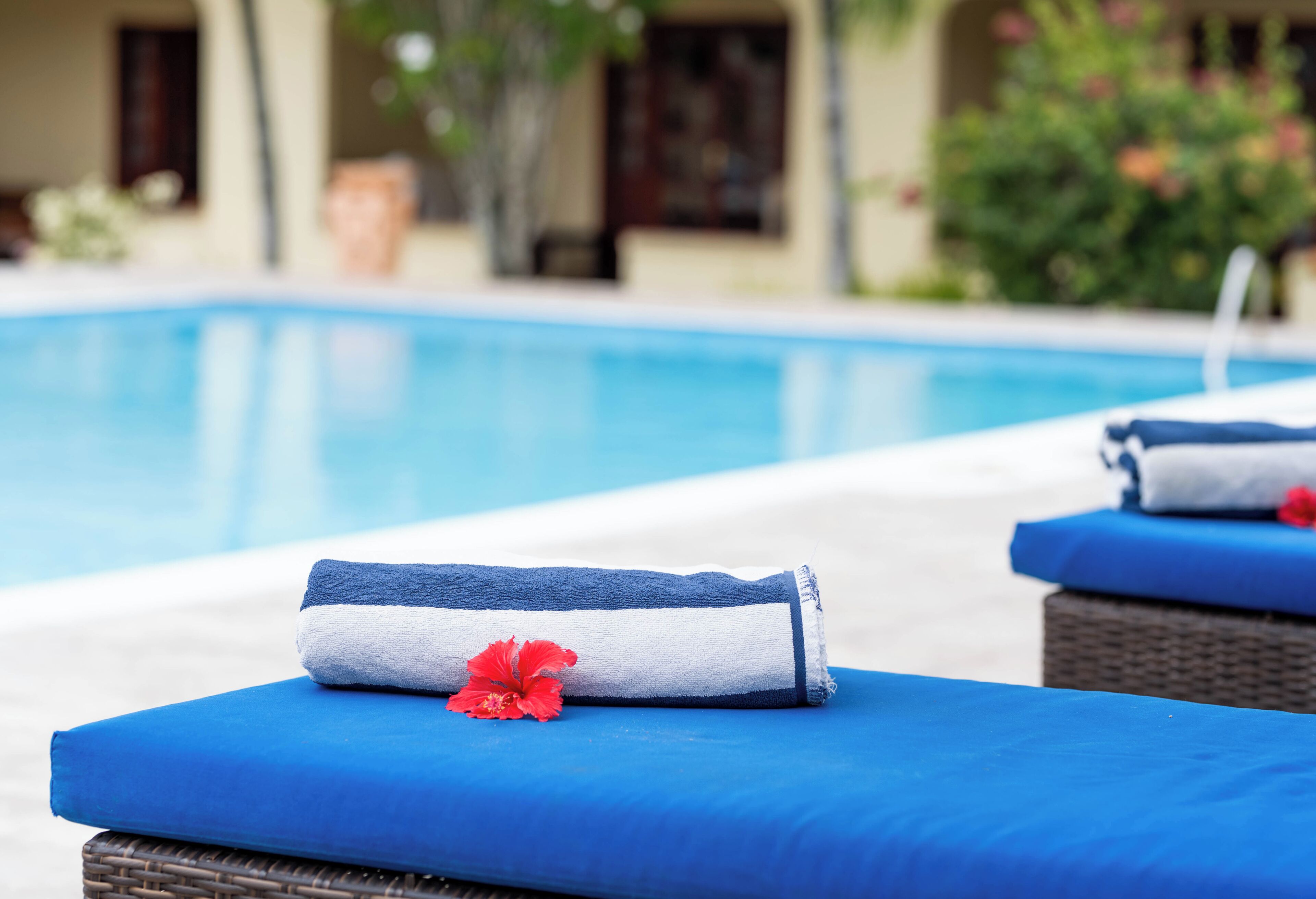 Zona de piscina con sillones reclinables y toallas, una piscina cristalina y un edificio con ventanas y vegetación al fondo.