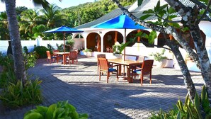 Un patio exterior con mesas y sillas bajo sombrillas azules, rodeado de plantas en macetas y una pared blanca.