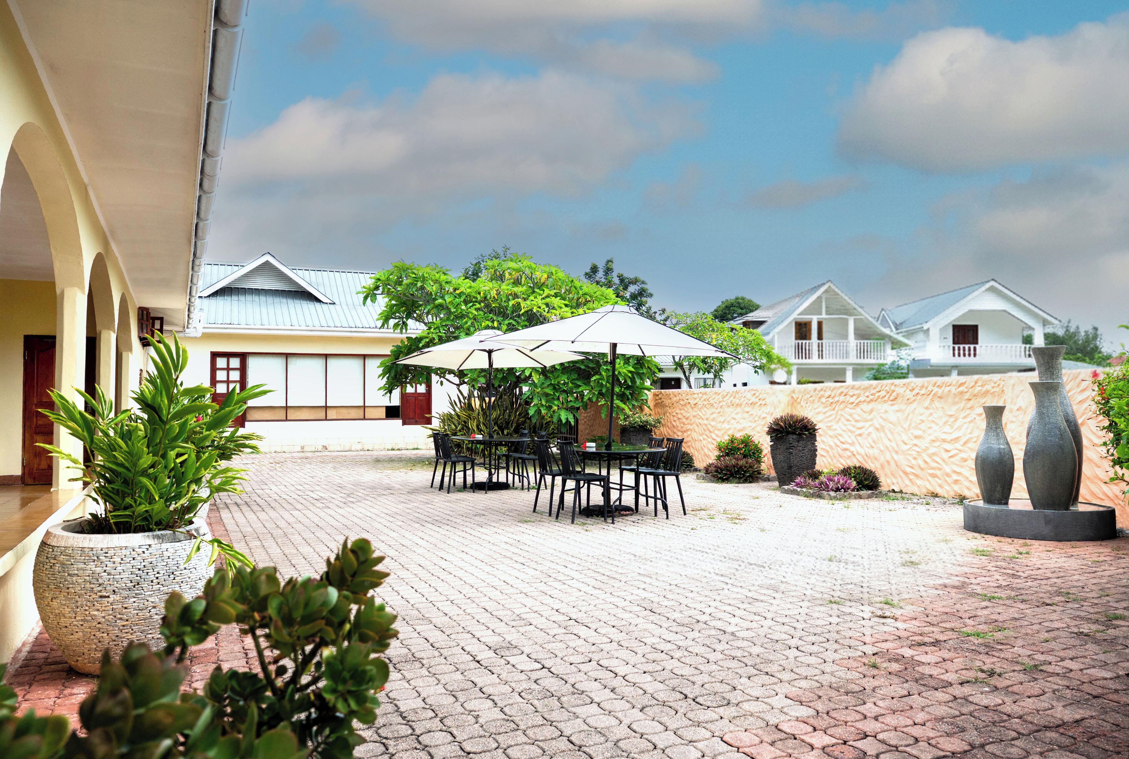A courtyard with a paved walkway, outdoor seating, potted plants, and a building with a sloped roof in the background.