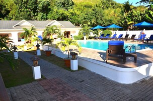Zona de piscina con sillas de descanso y sombrillas, rodeada de vegetación y un edificio con fachada blanca.