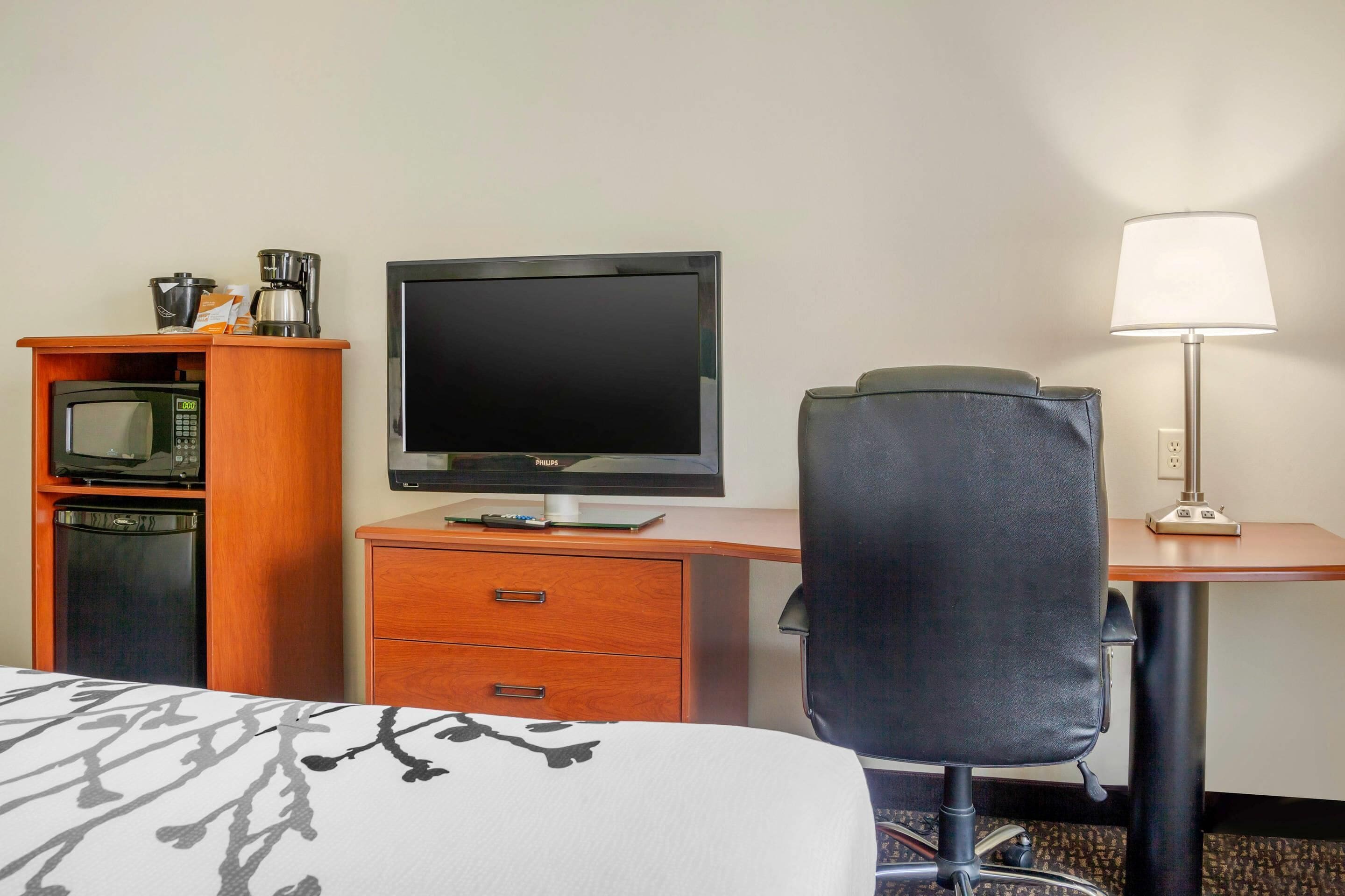 A hotel room with a bed, a desk with a computer monitor, a chair, a lamp, a microwave, and a coffee maker.