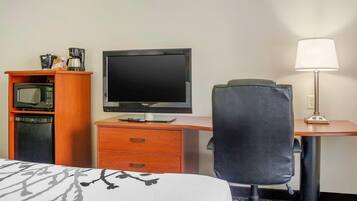 A hotel room with a bed, a desk with a computer monitor, a chair, a lamp, a microwave, and a coffee maker.