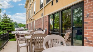 Patio esterno con barbecue, tavoli e sedie, accanto a un edificio con facciata in mattoni e ampie finestre.