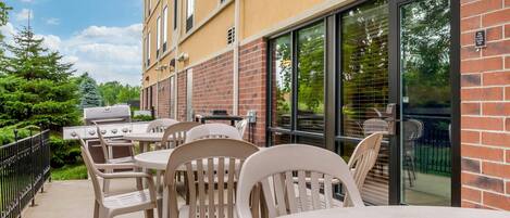 An outdoor patio with a grill, tables, and chairs, adjacent to a building with a brick facade and large windows.