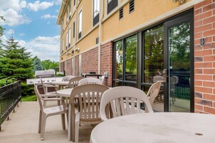 An outdoor patio with a grill, tables, and chairs, adjacent to a building with a brick facade and large windows.