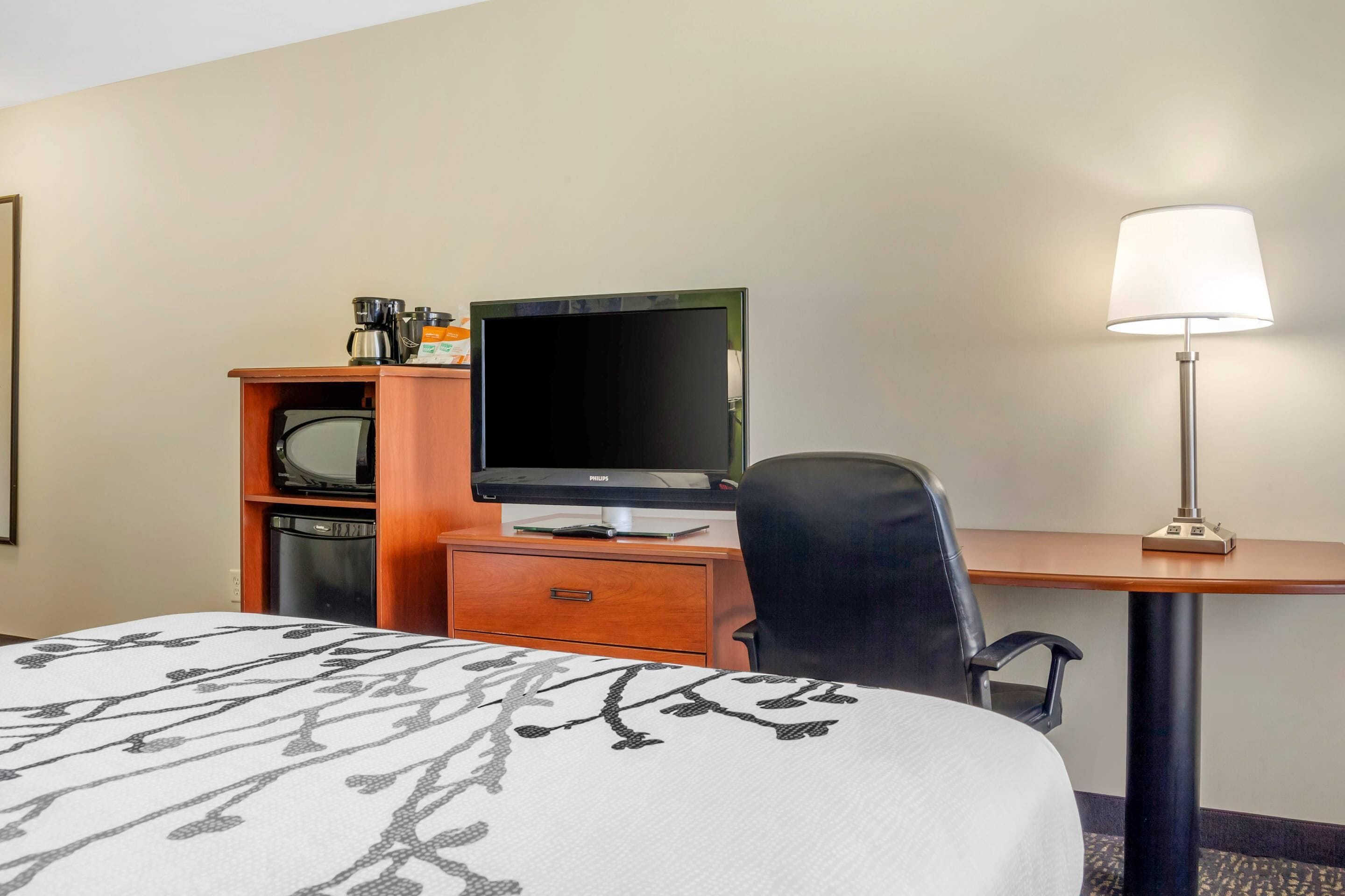 A hotel room with a bed, a desk with a computer and a lamp, a chair, and a television on a shelf.