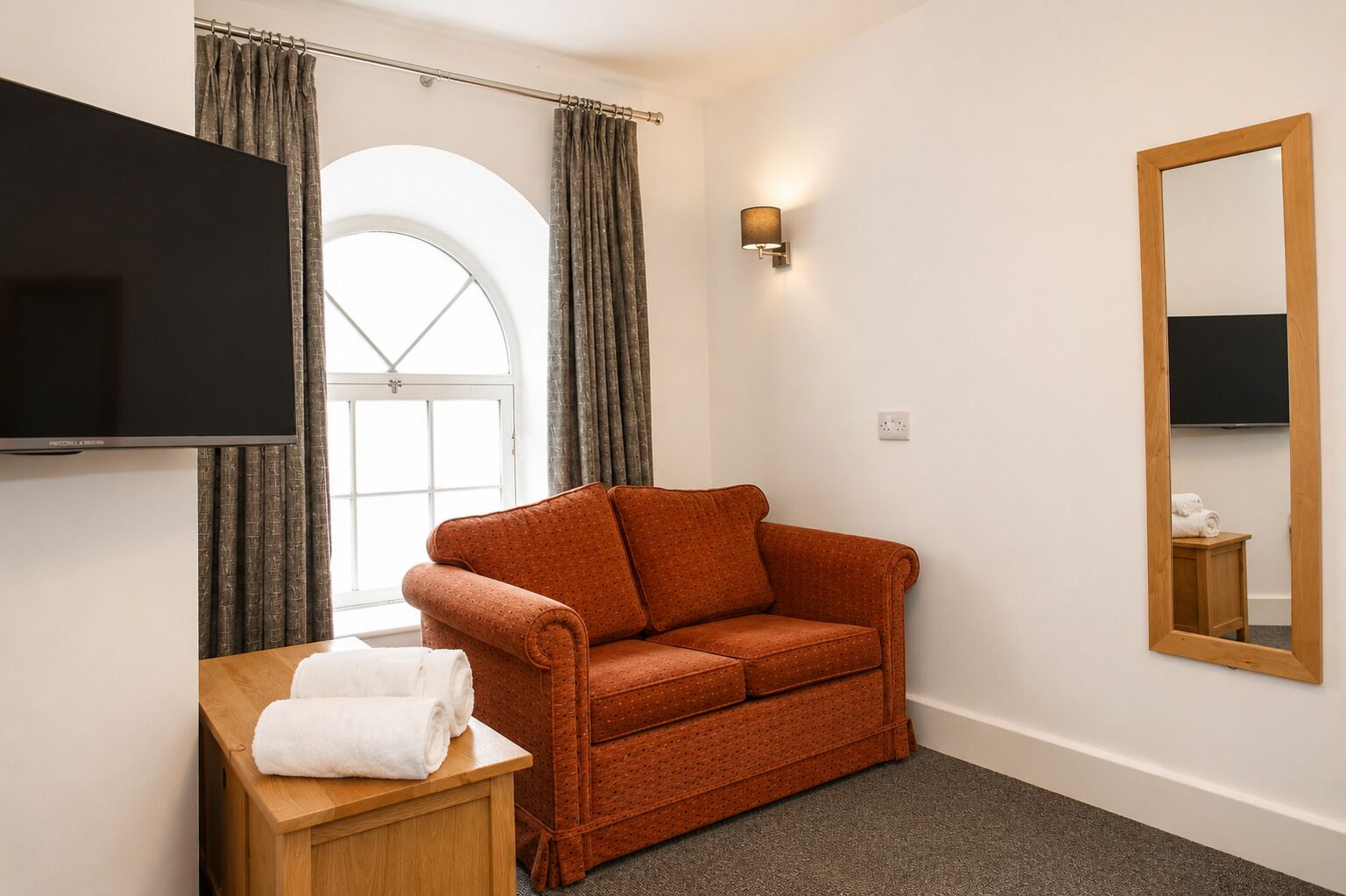 Une chambre d’hôtel avec un canapé, un téléviseur fixé au mur, une table d’appoint en bois avec des serviettes, un miroir et une fenêtre avec des rideaux.