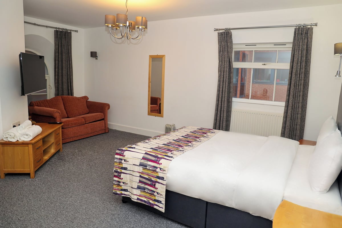 Habitación de hotel con cama, sofá, televisión, espejo y ventana con cortinas.