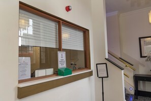 A reception area with a window, a sign for assistance, a small green box, and a framed notice on the wall. - Hotel 1843 Reading (Reading)