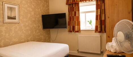 A hotel room with a bed, a flat-screen TV, a radiator, a window with curtains, a fan, and a remote control on a wooden table.