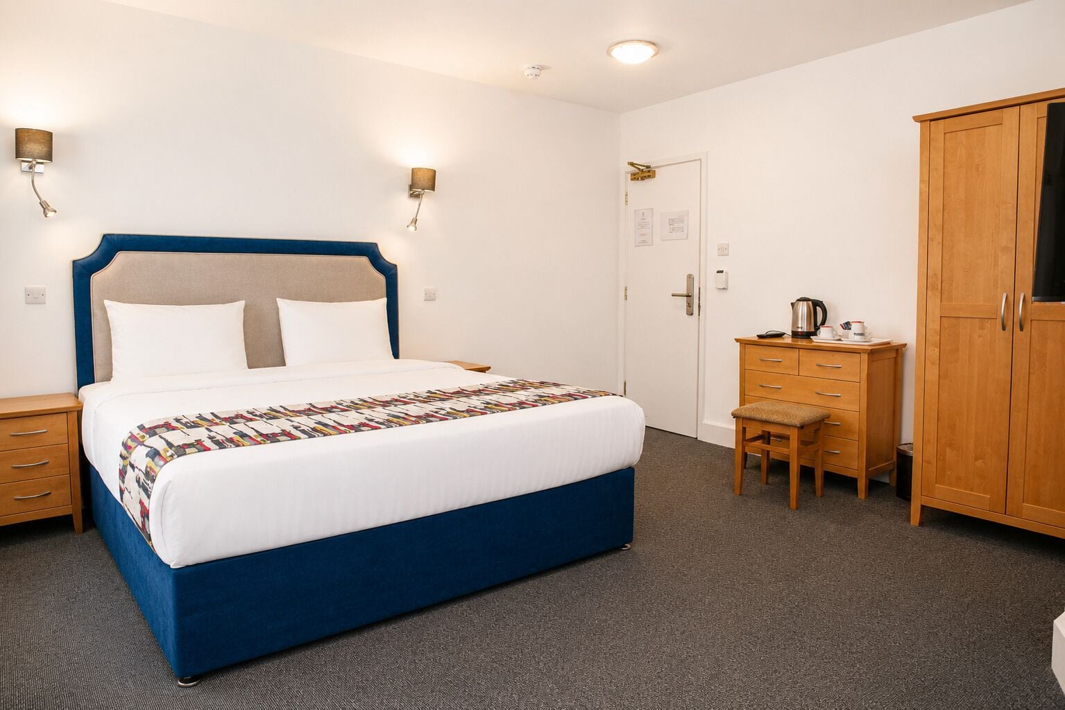 A hotel room with a bed, bedside tables, a desk, a chair, and a wardrobe.