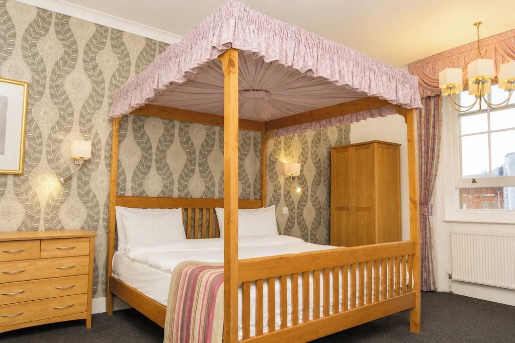 A four-poster bed with a canopy, a wooden dresser, a nightstand, and a window with curtains.