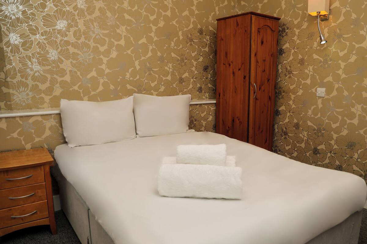Una cama bien hecha con sábanas blancas, una mesita de noche de madera con cajones y un armario de madera en una habitación con papel tapiz floral.