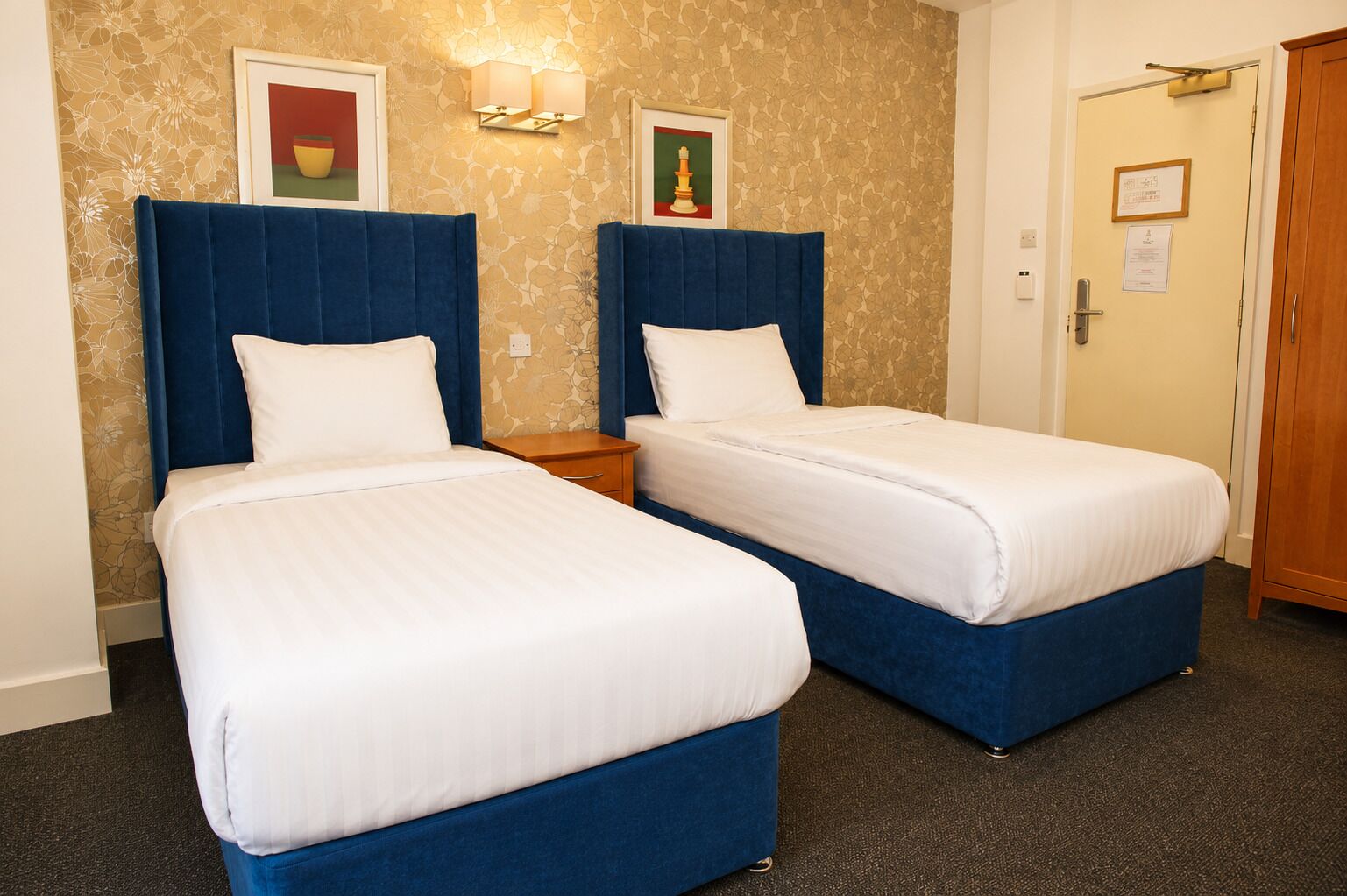 Habitación de hotel con dos camas, una mesita de noche, una lámpara de pared y una puerta con un letrero.