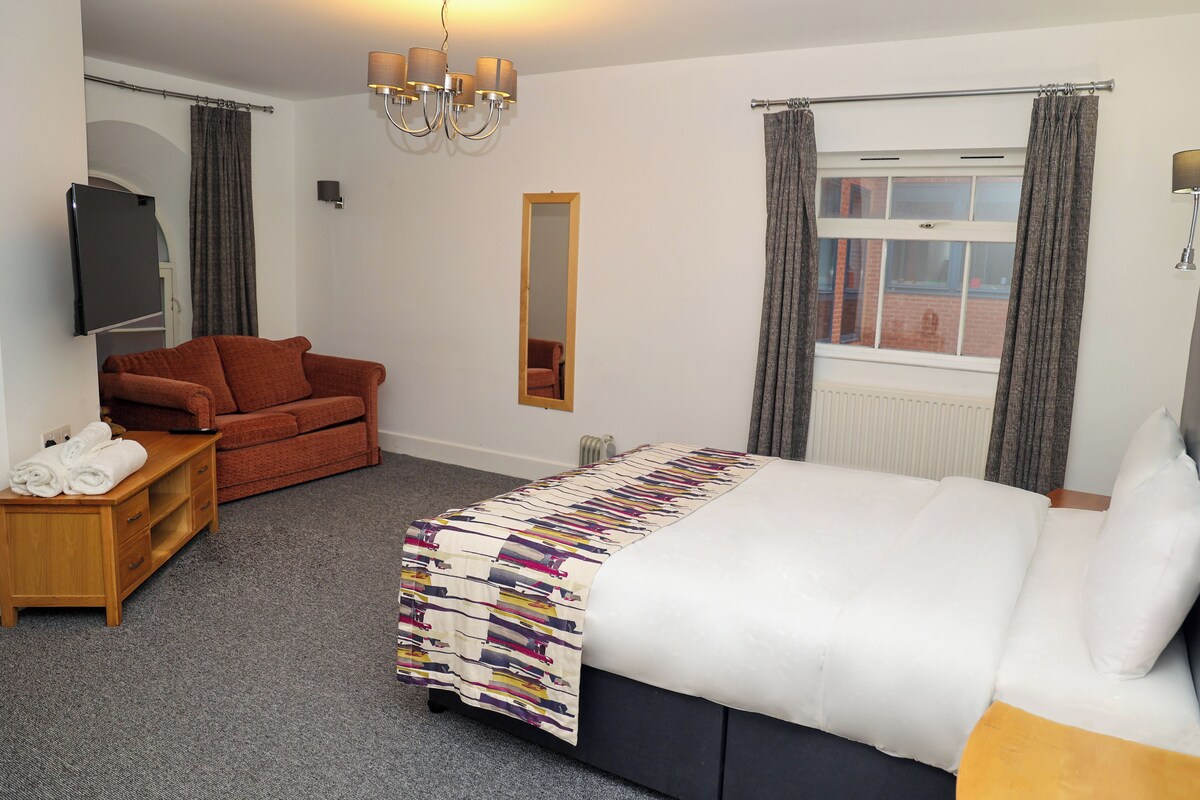 Habitación de hotel con cama, sofá, televisión, espejo y ventana con cortinas.
