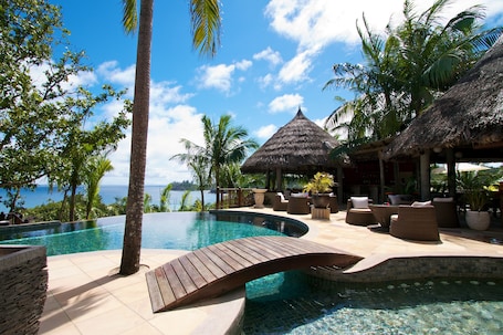 A pool area with a wooden deck, lounge chairs, and a thatched-roof structure.