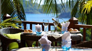 Una mesa de comedor dispuesta con una botella, copas y pan, sobre una mesa de mimbre con vista al mar.