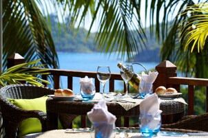 Una mesa de comedor dispuesta con una botella, copas y pan, sobre una mesa de mimbre con vista al mar.