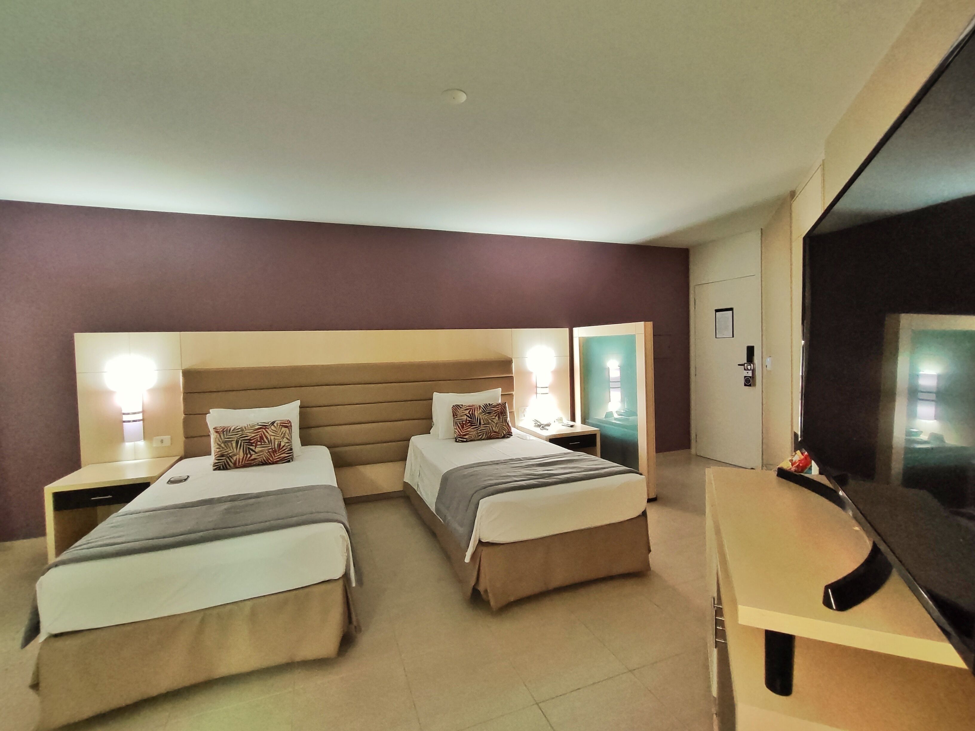 A hotel room with two beds, each with a headboard and bedside tables. There is a desk and a chair, and a television on the wall.