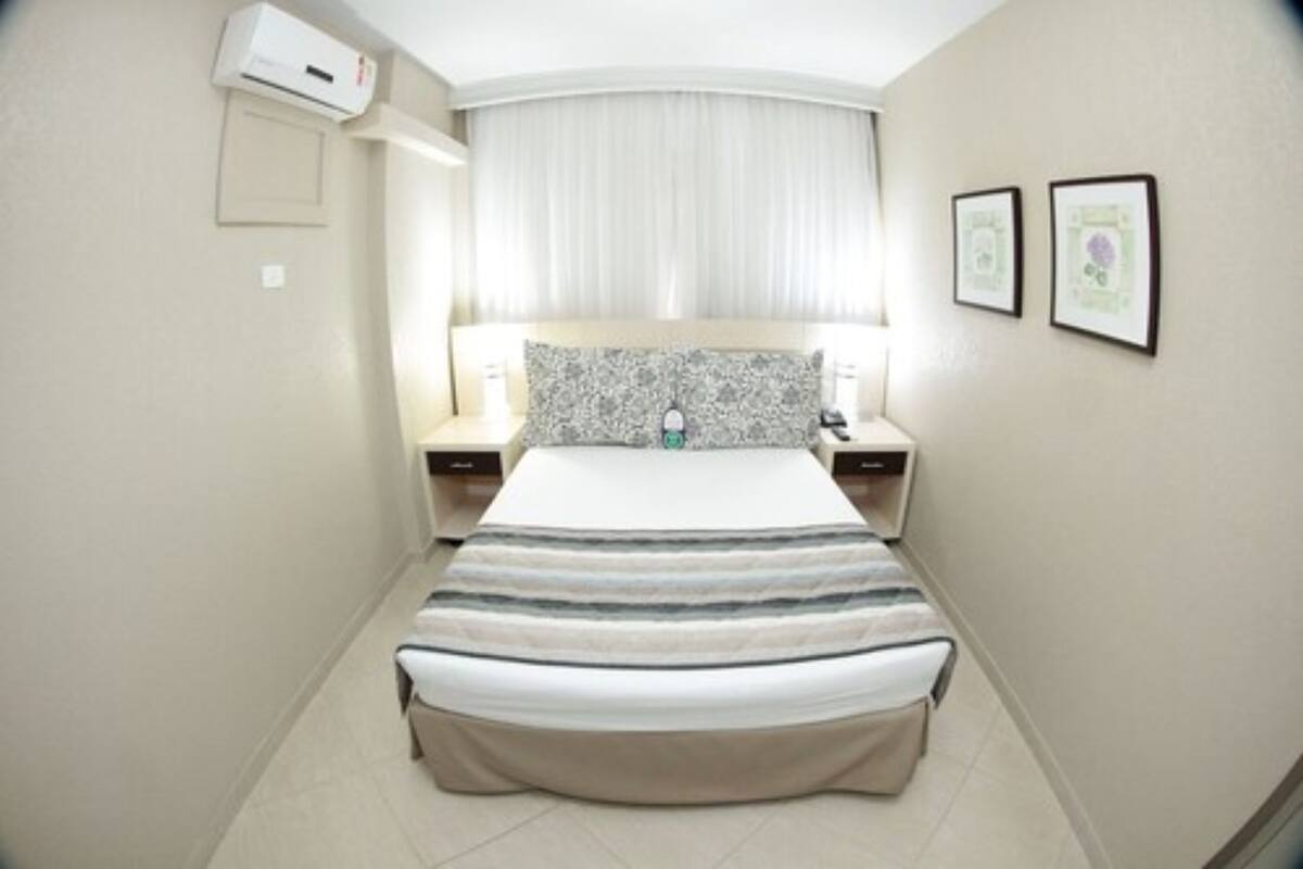 A hotel room with a bed, bedside tables, an air conditioning unit, a window with curtains, and two framed pictures on the wall.