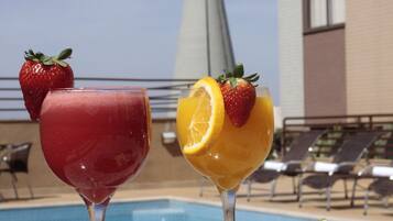 Mesa à beira da piscina com duas bebidas, um prato de queijos, azeitonas e um tabuleiro de presunto.