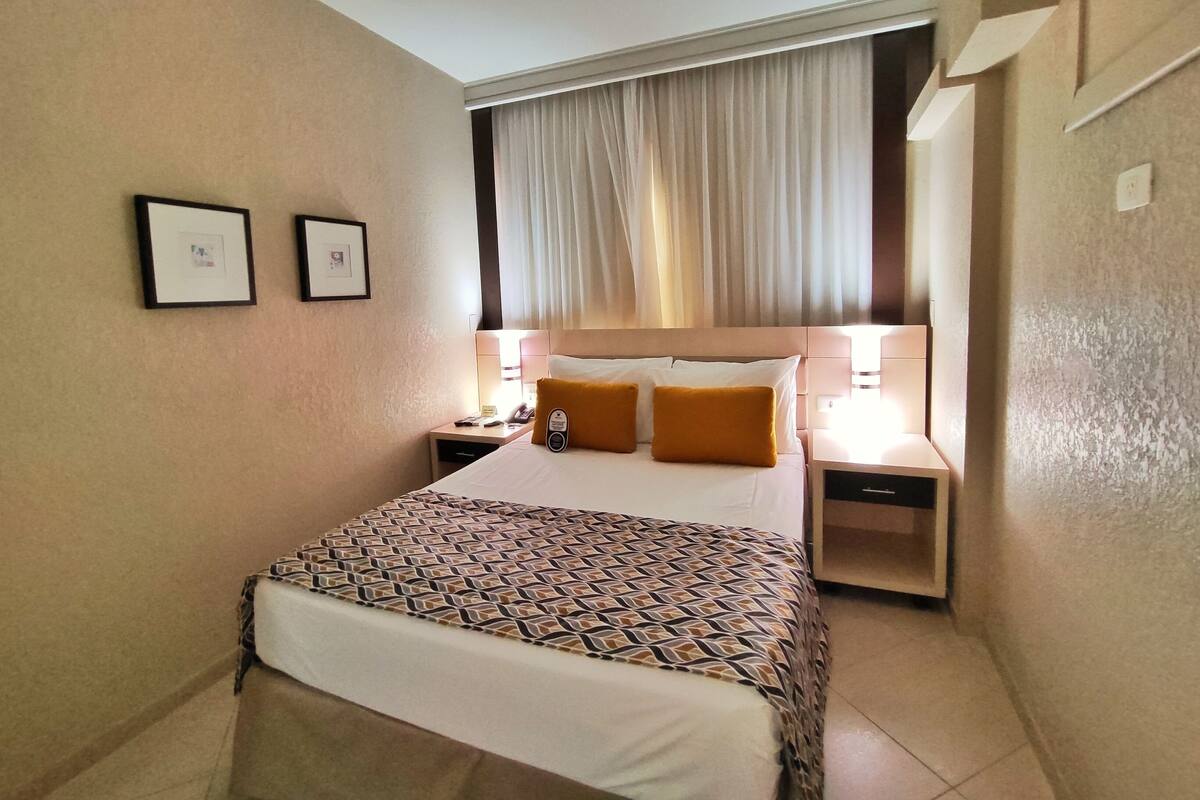 A hotel room with a large bed, two bedside tables, a nightstand, a window with curtains, and two framed pictures on the wall.