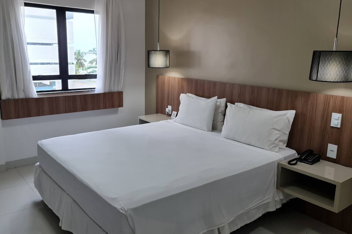 A hotel room with a large bed, two bedside tables, a nightstand with a phone, a window with curtains, and a wall-mounted lamp.