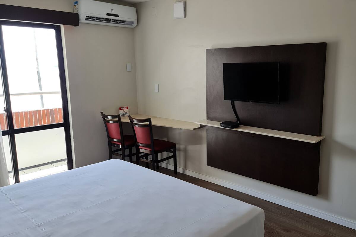A hotel room with a bed, a desk with two chairs, a mounted TV, and an air conditioning unit.