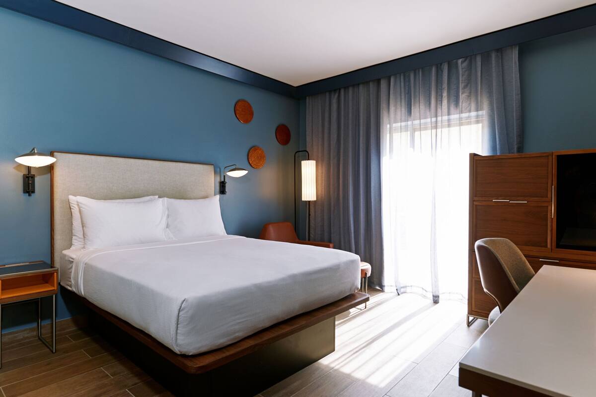 Habitación de hotel con una cama grande, un escritorio, una silla, un televisor y una ventana con cortinas.