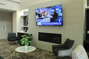 A modern hotel lobby with a flat-screen TV displaying a news broadcast, comfortable seating, a small table with a vase, and a fireplace. -  La Quinta Inn and Suites by Wyndham Schertz-San Antonio-Selma (Schertz)