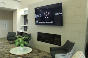 Un vestíbulo moderno con un televisor de pantalla plana mostrando 'Queen II Our Now', una chimenea, asientos cómodos y una alfombra estampada.
