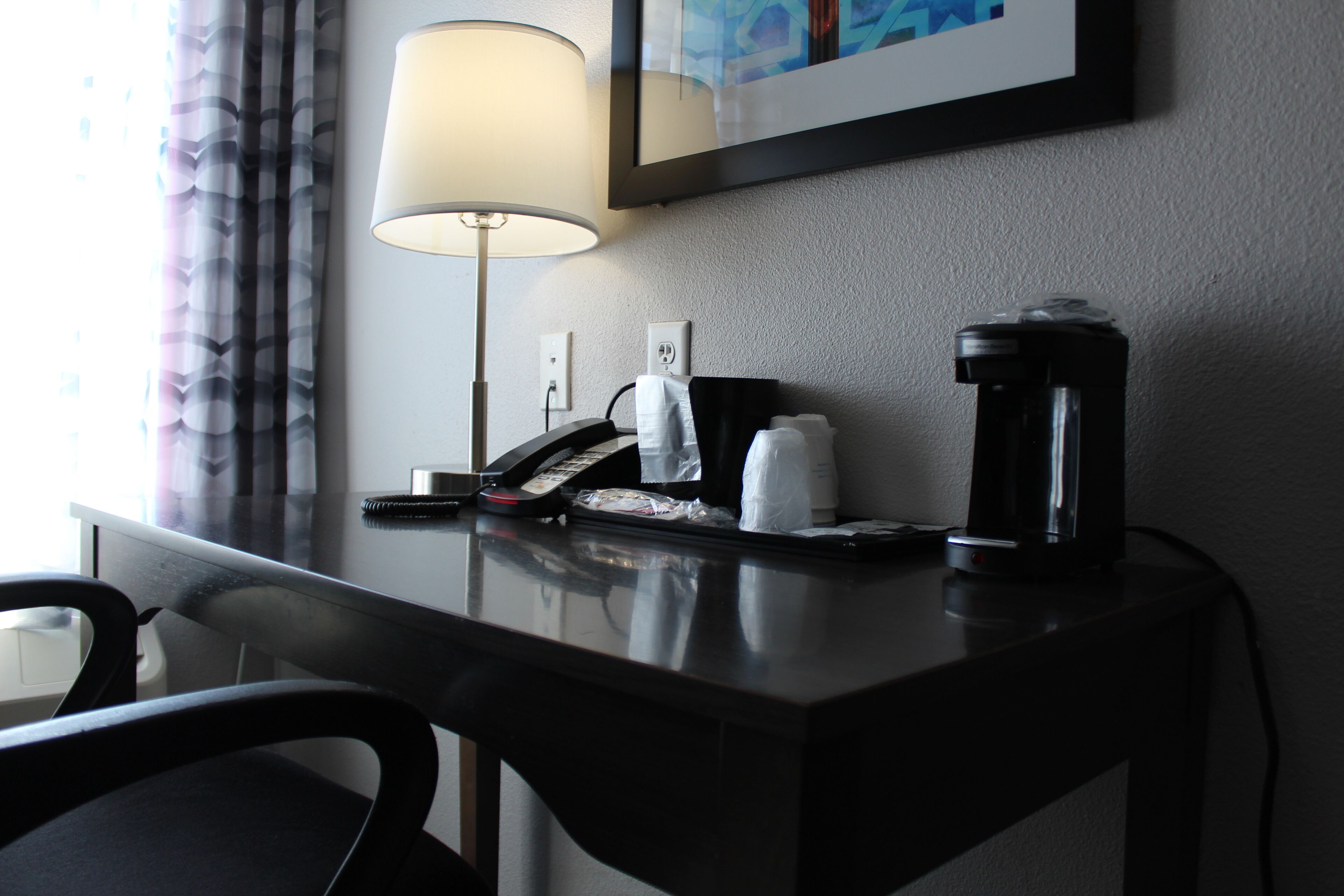 Escritorio de hotel con lámpara, teléfono, cafetera y un cuadro enmarcado en la pared.